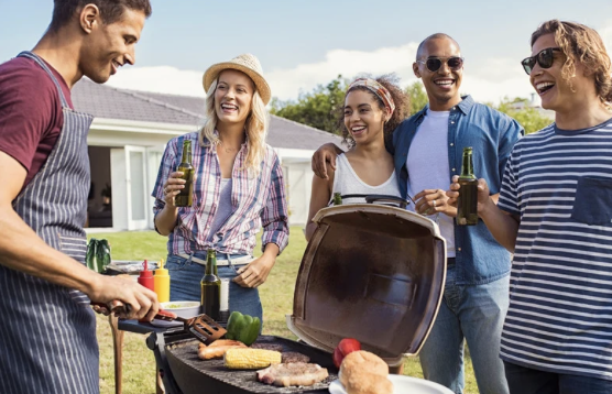 Family hosting a barbecue, all gathered around a grill while a man cooks.